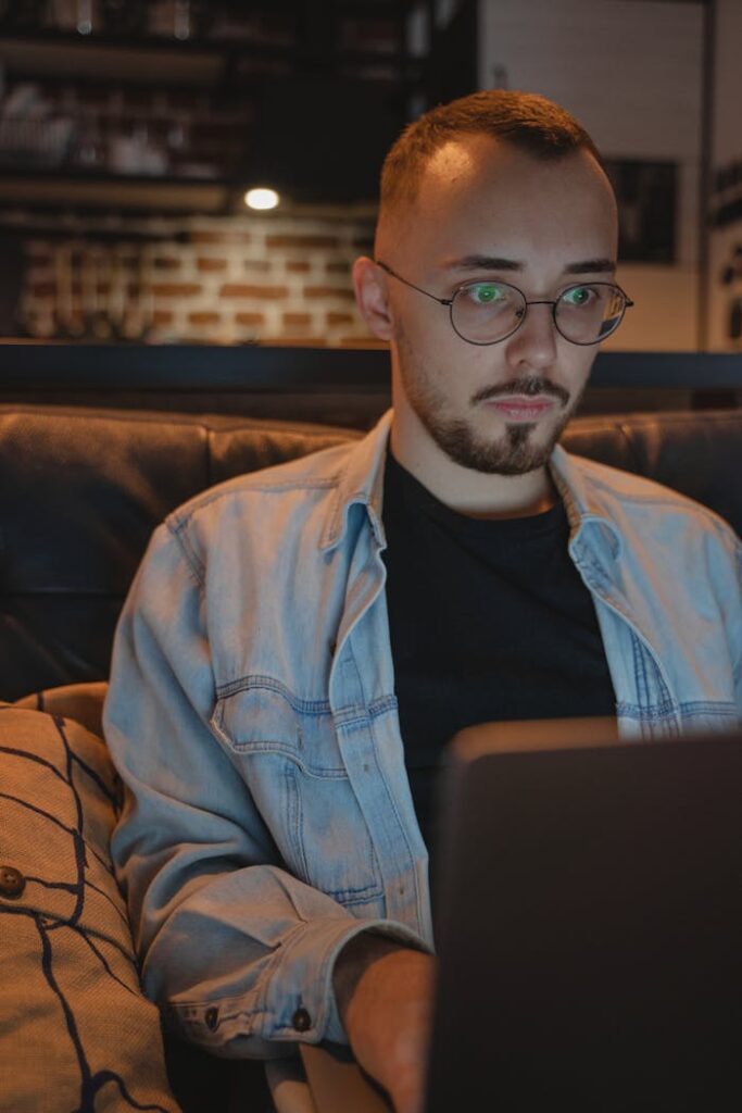 A young man with glasses works intently on his laptop in a cozy, indoor setting.